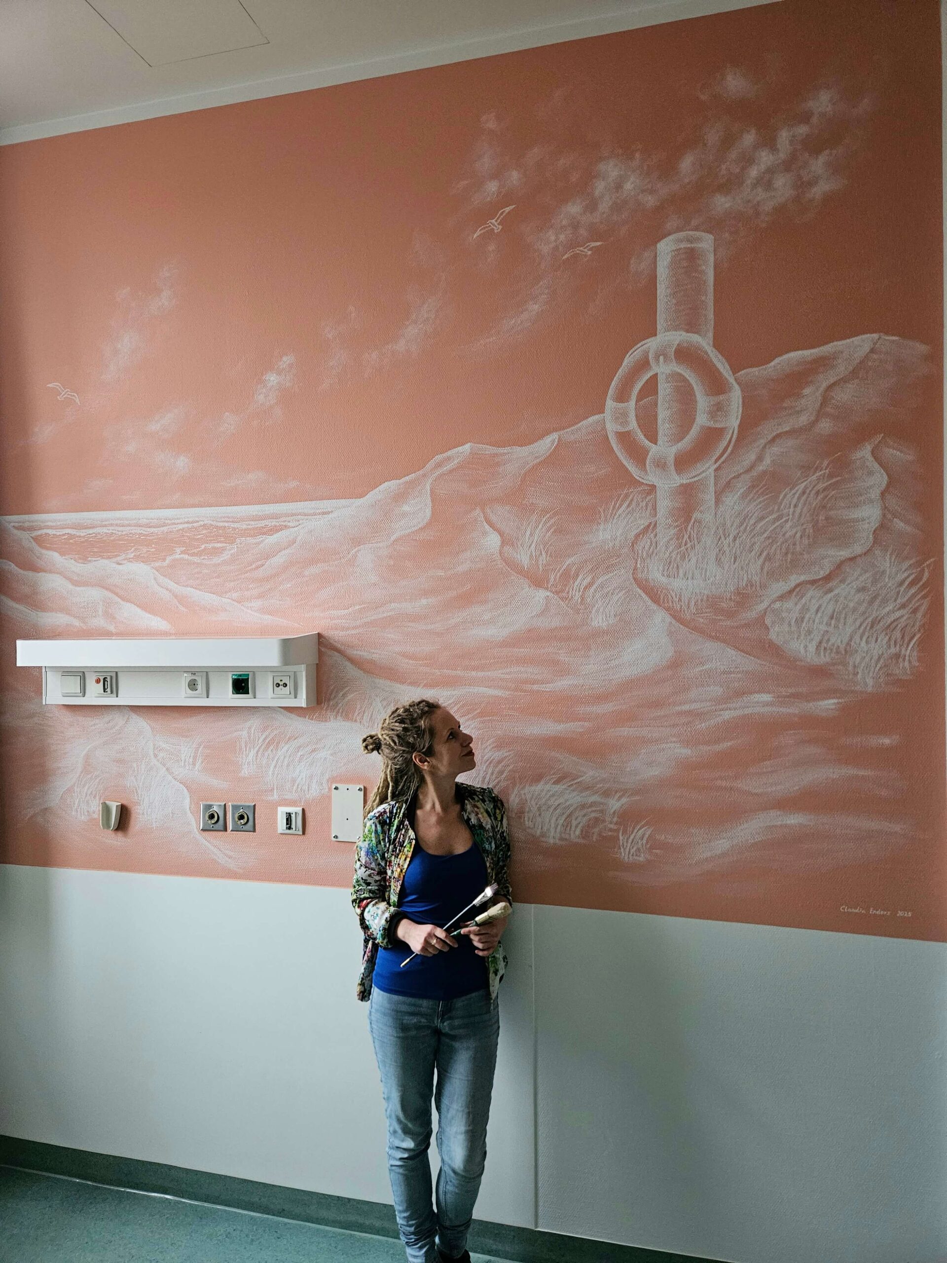 Lachsfarbene Wand mit einer Wandmalerei, auf der ein Rettungsring und eine Dünenlandschaft zu sehen sind.