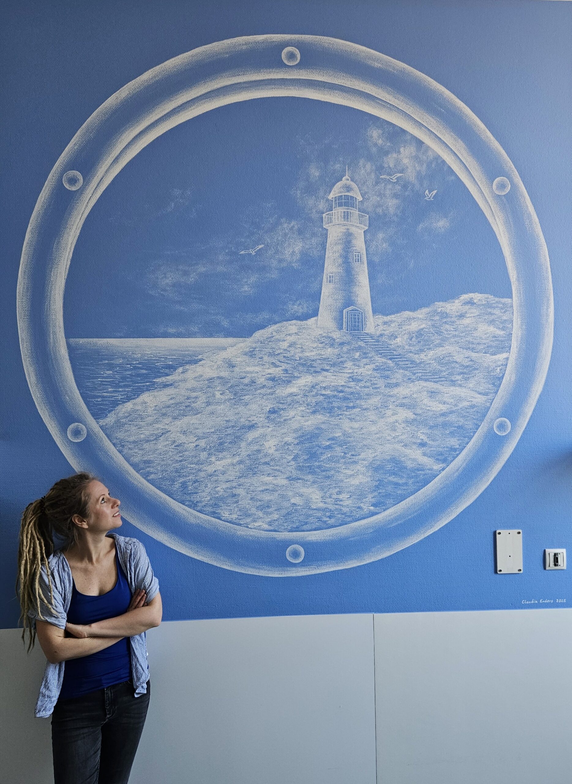 Blaue Wand mit einem Wandgemälde in Weiß. Das Motiv zeigt ein Bullauge, durch das man auf eine Klippe mit Leuchtturm schaut.