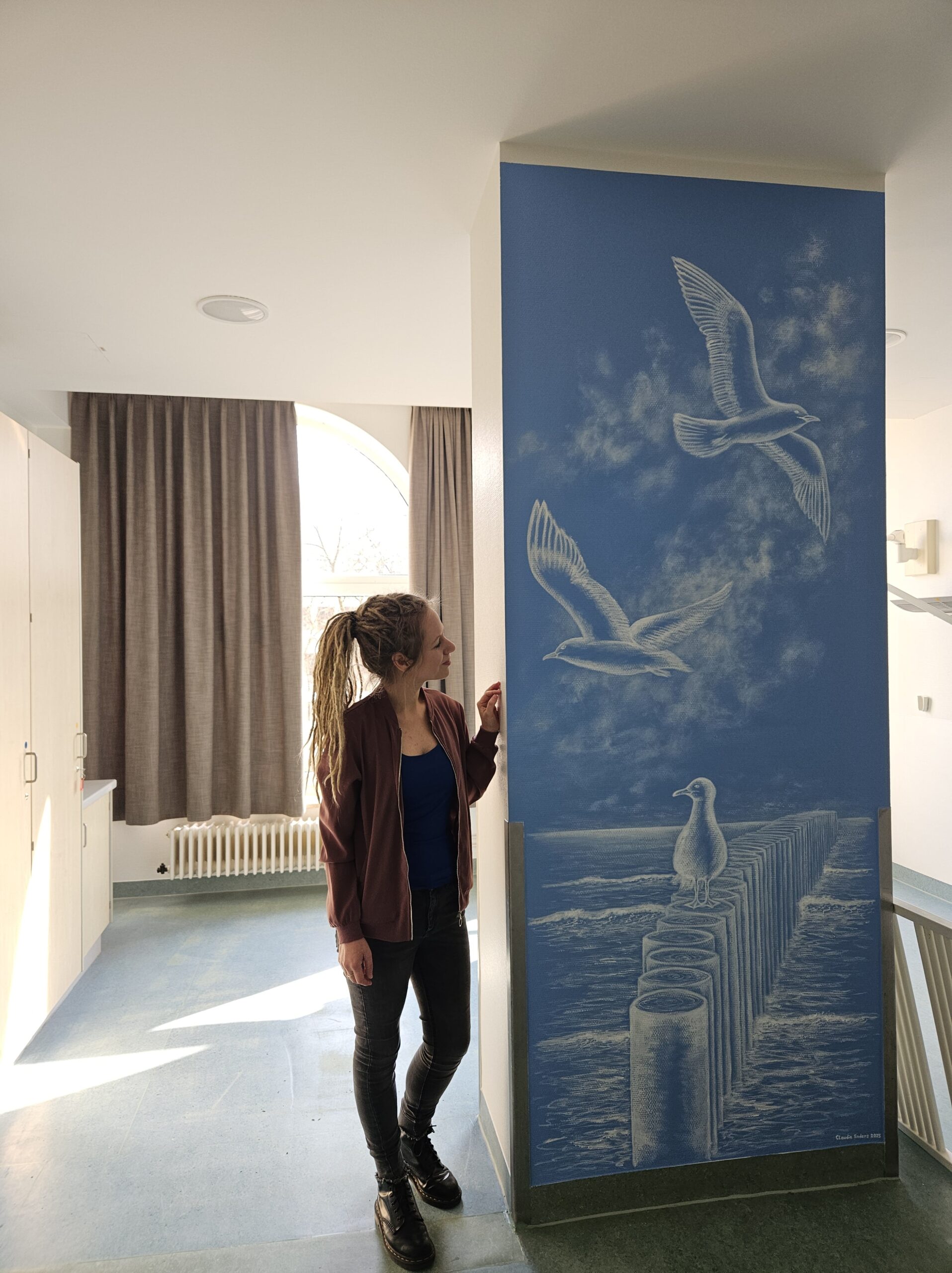 Blaue Wand mit einem Wandgemälde in Weiß. Das Motiv zeigt Möwen und einen Wellenbrecher im Meer.