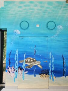 Zu sehen ist eine Detailaufnahme eines Wandgemäldes: Man sieht eine Schildkröte, die durch Seegras schwimmt.