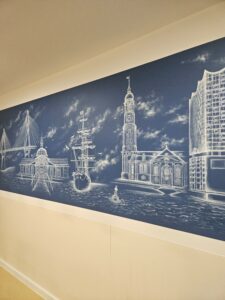 Detaillierte Malerei auf blauem Untergrund. Dargestellt sind Sehenswürdigkeiten der Stadt Hamburg. Die Wandmalerei ziert einen Aufenthaltsraum im Agaplesion-Krankenhaus in der Hansestadt.