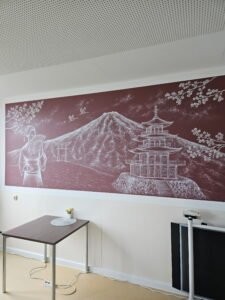 Japanisch inspiriertes Wandgemälde auf der geriatrischen Station des Agaplesion Diakonieklinikums Hamburg. Dargestellt sind eine Geisha, Mandschurenkraniche, ein Torii-Tor, der Berg Fuji und eine Pagode. Das Wandbild wurde im Zuge der Neugestaltung der Station geschaffen und trägt zu einer ruhigen, freundlichen Atmosphäre für Patientinnen und Patienten, Angehörige sowie das Klinikteam bei.
